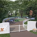 Kids Playground With White Fence Circular Path Tire Log Stumps Green Trees And Birdhouse