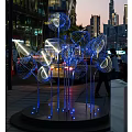Illuminated Outdoor Sculpture with Blue Lights and Metal Flower Design in Urban Night Background