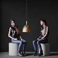 Two women sitting chatting on white cylindrical stools with gold pendant light against dark background