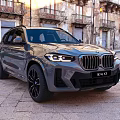 Gray BMW X3 Parked On Cobblestone Square With Historic Buildings And Headlights On