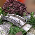 Stone Arch Bridge Surrounded By Red Shrubs Green Plants And Pebble Ground With Sunset Glow