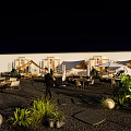 Outdoor Patio Night View With Wooden Tables Chairs Green Trees And String Lights
