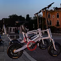 White Folding Electric Motorcycle Parked In Evening Stone Square With Buildings And Pedestrians