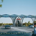 Modern Architectural Entrance Gate With Arched Metal Framework And Surrounded by Vehicles Pedestrians Green Trees