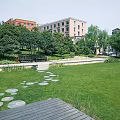 Park Landscape Featuring Green Lawn Circular Stone Stepping Stones Wooden Deck Bench Trees And Buildings