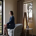 Businesswoman Sitting On Blue Armchair By Window With Curtains Floor Lamp And Outside Trees
