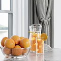 Glass Pitcher With Orange Drink And Bowl Of Oranges By Window Gray Curtains