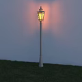 Outdoor Street Lamp with White Post Illuminated Warm Yellow Light on Grassy Ground Against Wall