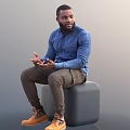 Man In Blue Denim Shirt Brown Pants Sitting On Gray Cube Gesturing With Hands