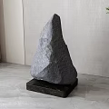 Gray Stone Sculpture On Black Wooden Base With Light Wall Curtain And Green Plant