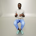 Man sitting gesturing in light gray shirt blue pants light blue shoes simple gray background