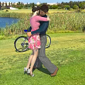 Couple Hugging Outdoors On Green Grassland With Lake And Bicycle
