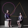 Two Women Sitting on White Cubes Holding Pink Folders with Pink Circles and Yellow Triangles