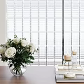 White Roses in Glass Vase with Green Leaves on Wooden Table by Window Blinds