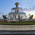 Garden Ornament Feature With Multi Tiered Fountain And Swan Sculptures Surrounding Flower Bed At Sunset