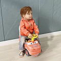 Boy in Orange Jacket Sitting on Yellow Toy Car Against Gray Wall and Light Floor