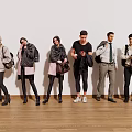 Group of people in various outfits standing on wooden floor with white wall holding bags