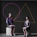 Two Women Sitting on White Cubes Holding Pink Folders with Pink Circles and Yellow Triangles