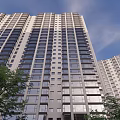 Modern Tall High Rise Apartment Building With Evening Window Lights And Green Trees