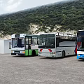Different Colored Buses Parked Outdoor With Green Hills And White Building
