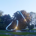 Curved Stainless Steel Sculpture in Outdoor Park with Green Trees and Shining Light
