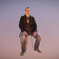 Man sitting on chair wearing black leather jacket brown t-shirt ripped blue jeans gray shoes