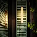 Modern Wall Sconces With Warm Glowing Light Against Green Louvered Shutters And Foliage
