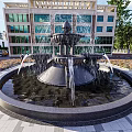 Ornate Fountain With Central Sculpture Water Flowing In Urban Plaza With Buildings And Greenery