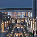 Bustling Evening Commercial Street with Shoppers Stores Hanging Lights Glass Facades and Green Plants